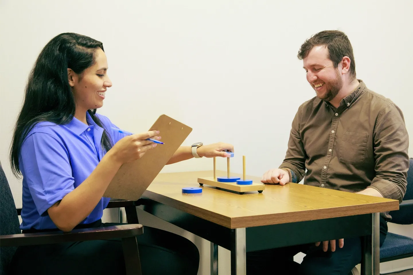 A female research assistant sits across from a male study participant, working on cognitive memory exercises using blue circles and wooden pegs.