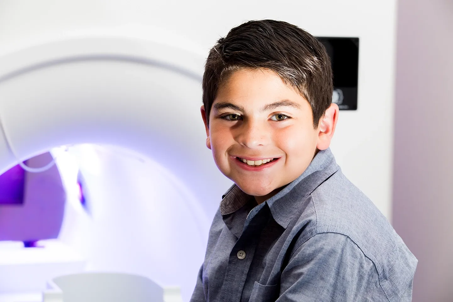 A young boy is smiling with an MRI scanner in the background.